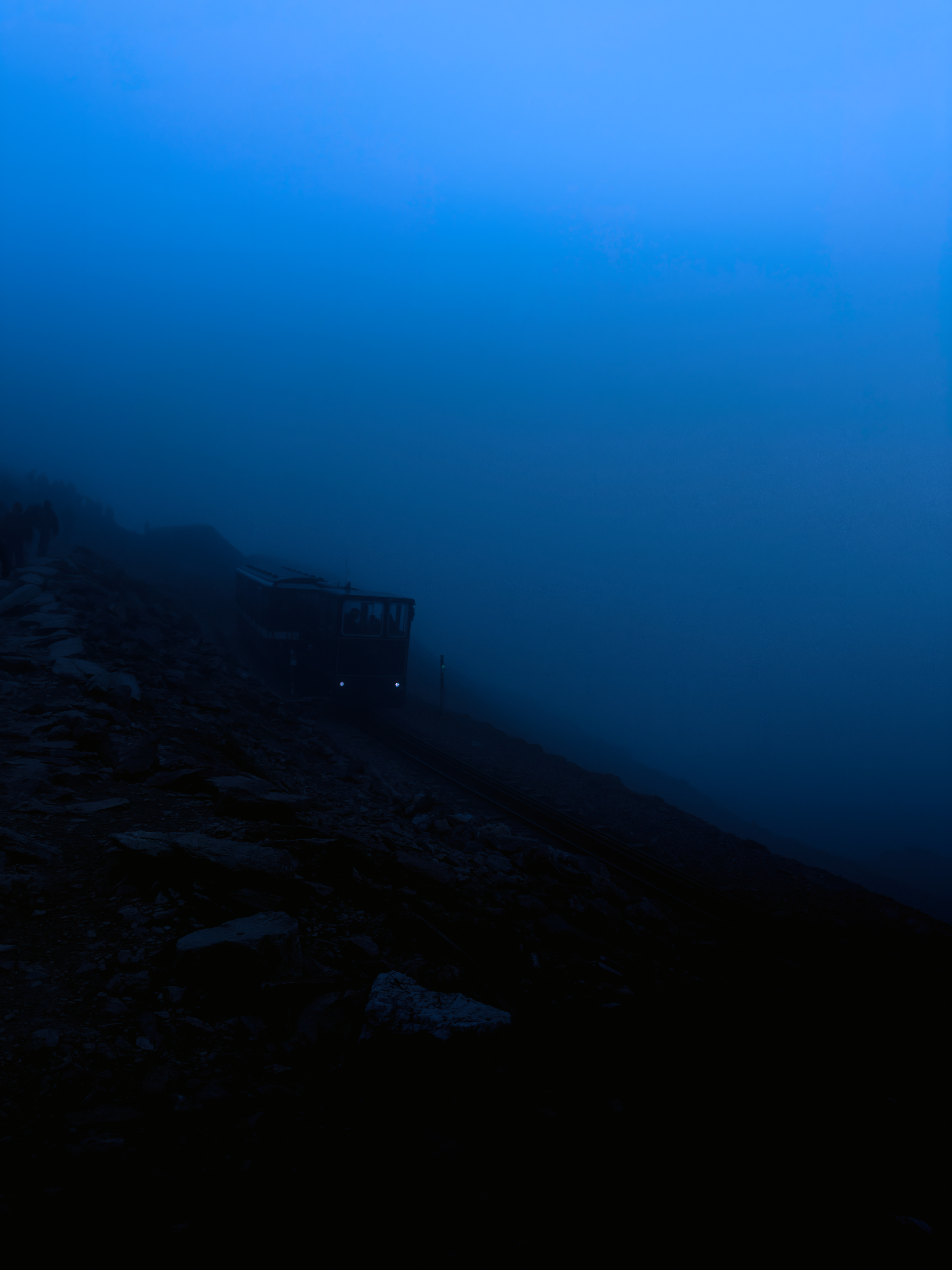 A mountain train emerging through deep blue mist on a rocky slope of Yr Wyddfa